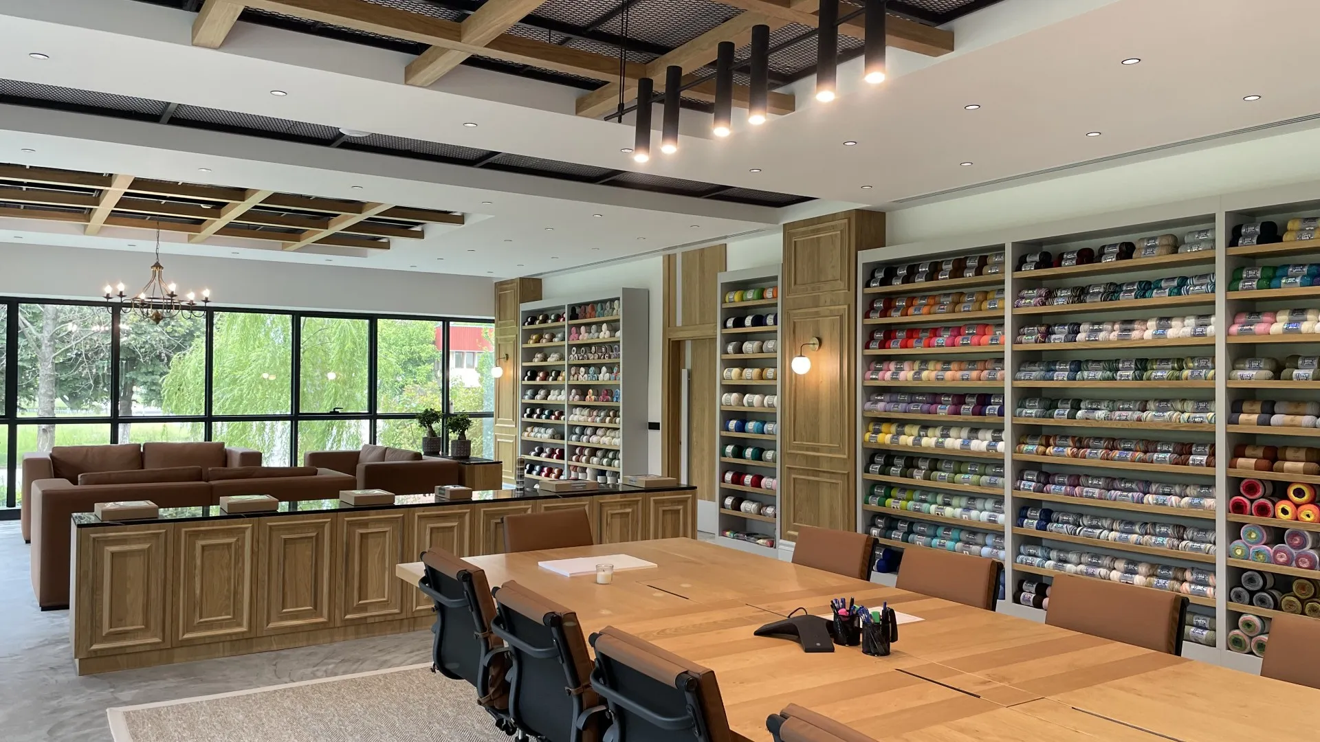 Modern showroom with wooden furniture, large meeting table, leather chairs, and extensive wall shelving displaying colorful yarn samples organized by color, large windows with natural light