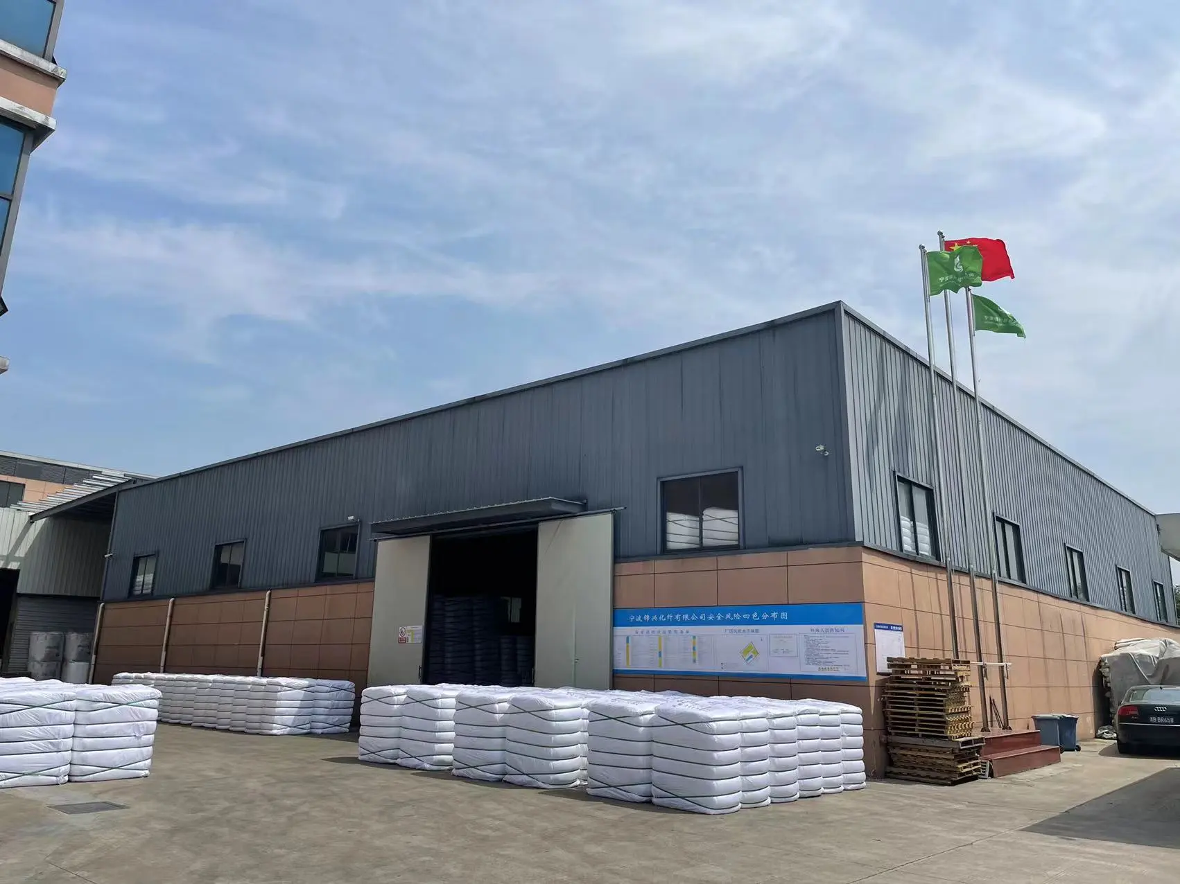Industrial factory building exterior with gray metal siding, brown base, and flags flying on the roof. Stacks of white materials visible outside.